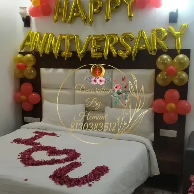 A romantic anniversary decoration right in the bedroom. The setup includes "Happy Anniversary" foil balloons above the headboard, simple balloon clusters, and "I Love You" written with rose petals on the bed.