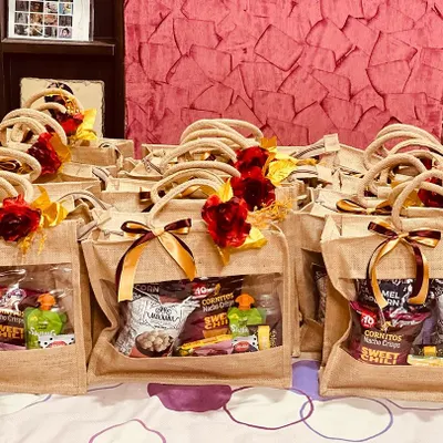 A large batch of wedding room hampers, ready to welcome guests. These eco-friendly jute bags are filled with snacks and drinks, and decorated with a red rose and ribbon.