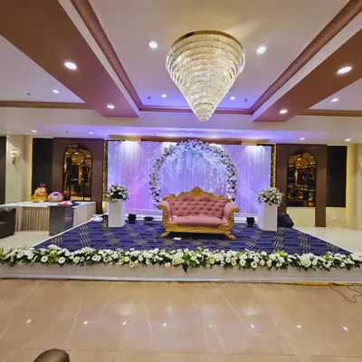A wide view of a stage designed for a ring ceremony in a banquet hall. The setup includes a floral arch, a pink and gold sofa, and a stage skirted with white flowers.