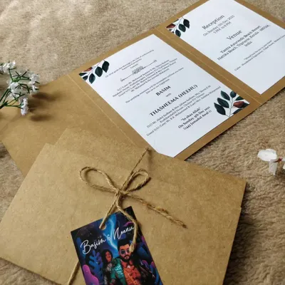 A rustic tri-fold invitation made from kraft paper, featuring simple leaf illustrations. The suite is tied with natural twine and includes a personalized tag with the couple's photo.