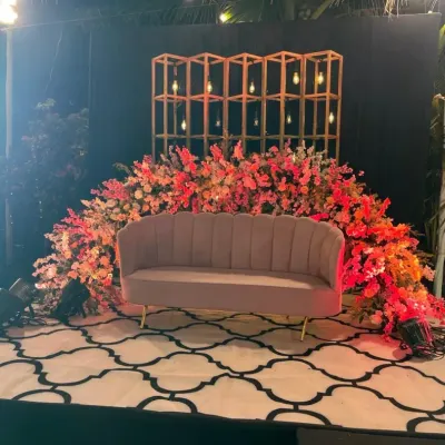 This photo booth combines rustic and modern elements. It features a geometric gold shelf with Edison bulbs, set against a backdrop of lush, colorful flowers and a patterned rug for a stylish photo opportunity.