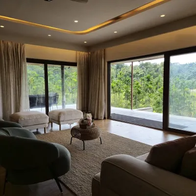 The living room of our "Songs of the Treepie" homestay in Coorg is designed to frame the stunning natural landscape. Expansive sliding glass doors open onto a terrace, blurring the lines between the comfortable interior and the verdant forest outside.