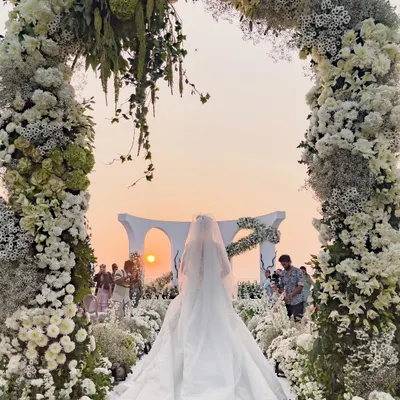 A fairytale moment as the bride walks towards a breathtaking sunset ceremony. The floral archway frames the scene, creating a perfect, romantic path for her grand entrance at this stunning white wedding in Goa.