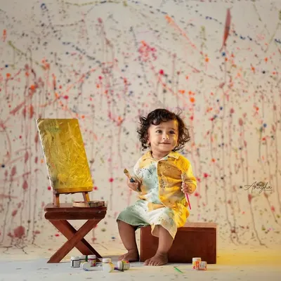 A toddler dressed as a little artist smiles proudly next to his painted canvas on an easel, against a paint-splattered wall.