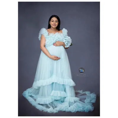 A full length portrait showcases the beauty of a flowing maternity gown. This mother to be looks radiant in a layered, light blue tulle dress, holding a simple bouquet that adds a soft, delicate touch to the image.