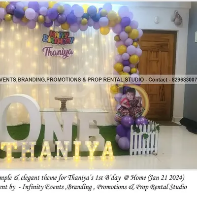 A simple and elegant theme for Thaniya's first birthday at home. This setup features a white curtain backdrop with fairy lights, a purple and gold balloon garland, and a "ONE" table, creating a magical corner.