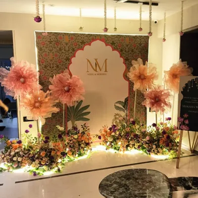 An elegant backdrop for an engagement or reception, featuring a custom monogram (NM) in a Mughal-style arch. The setup is decorated with large handmade paper flowers and fresh floral arrangements.