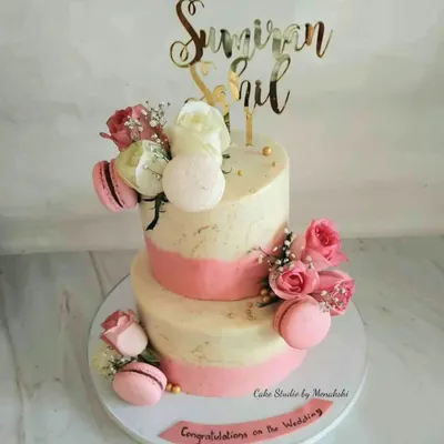 A beautiful two-tier wedding cake in white and pink. This no-fondant design is finished with buttercream and adorned with fresh roses and delicate macarons for a touch of elegance.