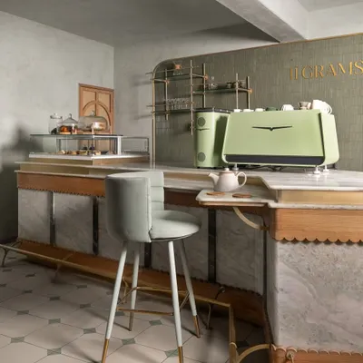 The main service counter at 11 Grams is crafted from white marble with delicate scalloped wood trim and brass details. The design ensures operational flow while serving as a beautiful centerpiece, complemented by a custom pale green espresso machine.