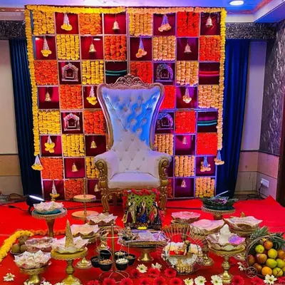 An elaborate Seemantha stage setup featuring an ornate high-back chair. The backdrop is a custom grid of fresh marigold and tuberose flowers, with traditional brass bells and a spread of offerings in front.