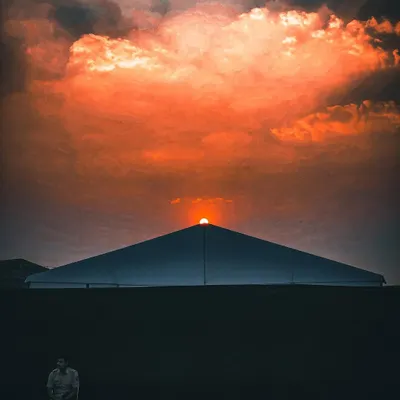 A minimalist and symmetrical composition of the sun setting behind a large tent, with a lone security guard seated, edited with a dramatic orange and black contrast.
