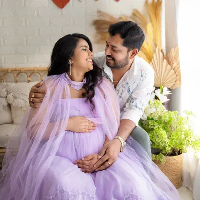 A happy, interactive moment between an expecting couple. The mother-to-be, in a lovely lavender gown, shares a smile with her partner in a bright, boho-chic studio setting.