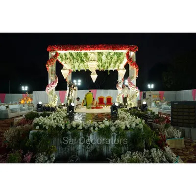 This is a large, open-air wedding mandap I set up for a night ceremony. The structure is decorated with red and white flowers, hanging chandeliers, and is surrounded by a garden of white flowers for a truly grand celebration.