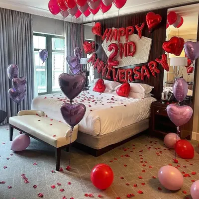 Celebrating a 2nd anniversary in style. This hotel room decoration uses a romantic mix of red and purple heart-shaped balloons, "Happy 2nd Anniversary" foil letters, and rose petals scattered across the floor and bed.