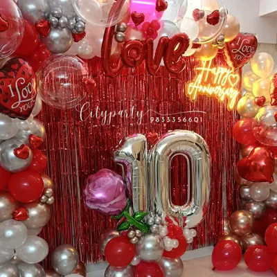 A complete view of our 10th-anniversary balloon decor package. It includes a full balloon arch, a red tinsel backdrop, large '10' number balloons, and a 'Happy Anniversary' neon sign.