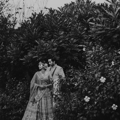 A classic black and white portrait of a couple in traditional Indian attire, standing amidst lush greenery. The timeless quality of the monochrome edit adds a layer of elegance and romance to the scene.