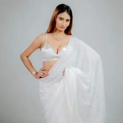 A full-length portrait against a clean grey background. The bright lighting beautifully illuminates the white satin saree, highlighting its texture and drape for a glamorous fashion shot.