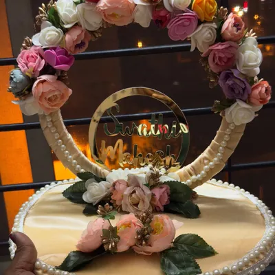 A close-up of a customized engagement ring platter. This design features a floral wreath, pearl detailing, and a golden acrylic cutout of the couple's names, Swathi and Mahesh.