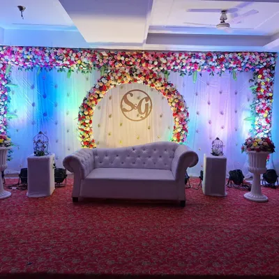 A complete stage view for an engagement, featuring a plush sofa for the couple. The backdrop includes a semi-circular floral arch and is lit with colorful spotlights to create a festive and romantic setting.