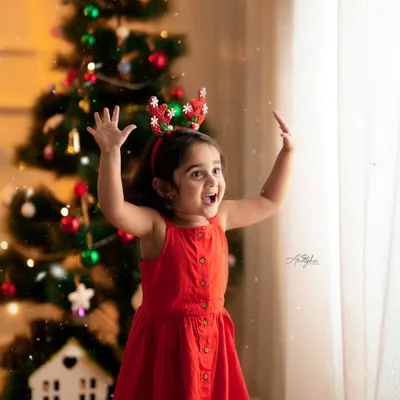 A young girl in a red dress and reindeer antlers raises her hands in excitement in front of a decorated Christmas tree.