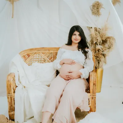 A full-length portrait from a boho-themed maternity session. The rattan furniture, pampas grass, and soft fabrics create a warm, earthy, and relaxed atmosphere.