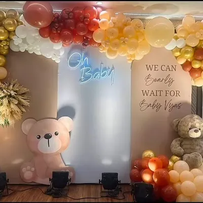 This teddy bear baby shower decor combines a warm color palette of red, gold, and peach with an "Oh Baby" neon sign. The setup includes two cute bear props and is personalized with the baby's name on the backdrop.