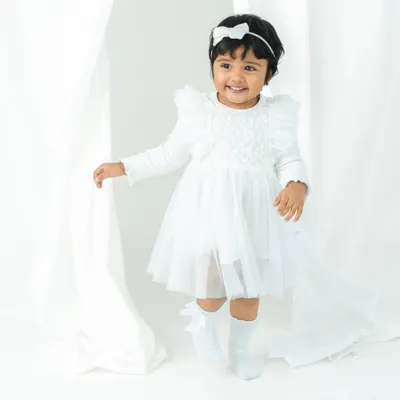 A vision in white. This two-year-old girl's bright smile and playful energy are captured beautifully against a simple, elegant white curtain backdrop.
