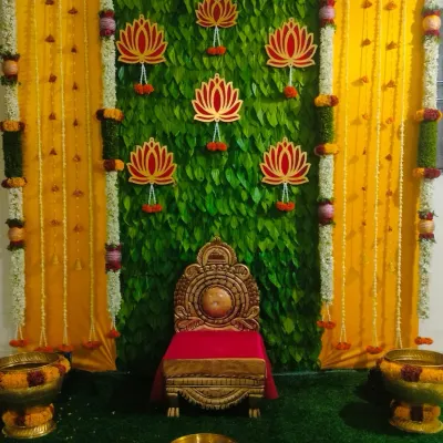 A beautiful setup for a half saree function. The backdrop is a wall of fresh betel leaves, accented with lotus cutouts and framed by yellow drapes and flower columns.
