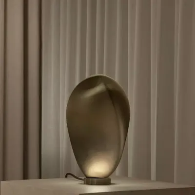 The Solace table lamp by Ross Gardam, with its visually enchanting teardrop shape, sits on a pedestal against a soft curtain. The bronze glass finish creates a warm, moody light, making it a perfect accent piece for a sophisticated interior.
