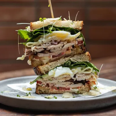 A towering club sandwich, stacked high with roasted chicken, bacon, egg, and fresh greens between slices of our toasted sourdough bread. This is a proper meal.