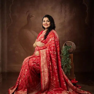 A regal portrait in a stunning red Banarasi saree. The backdrop, featuring a subtle peacock motif, adds a touch of traditional Indian artistry to this maternity photo.