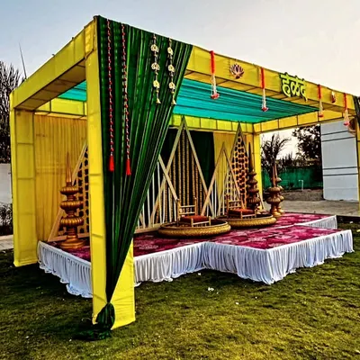 A full view of a beautiful outdoor Haldi mandap setup. The structure is decorated with yellow and green drapes, traditional brass lamps (diya stands), and comfortable seating for the main ceremony.