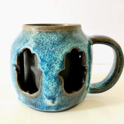 A full view of the double-walled Hamsa mug, showing the protective symbol carved on all sides. The beautiful blue glaze flows around the carvings, creating a piece that feels wonderful to hold and use.