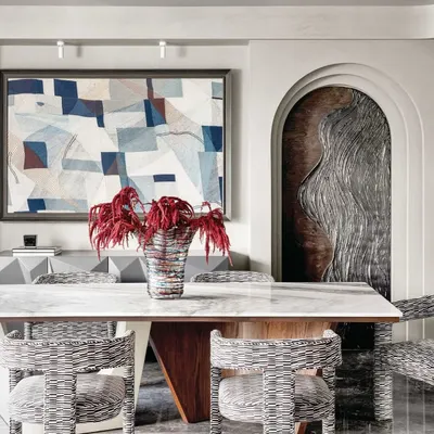 This dining space is a study in texture and form. The woven chairs, marble tabletop, and abstract art create a sophisticated yet inviting atmosphere. The arched doorway reveals a custom art panel, adding another layer of craftsmanship to the room.