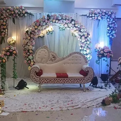 A classic engagement stage setup featuring an ornate white diwan sofa for the couple. The backdrop is a full circular arch made of dense pastel pink and white roses, complemented by matching floral stands and soft lighting.