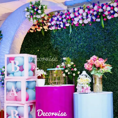 This modern baby shower decor combines several popular elements. It features a lush artificial green wall, a pastel blue arch, and clear acrylic boxes spelling "BABY" filled with pink and blue balloons for a gender-neutral theme.