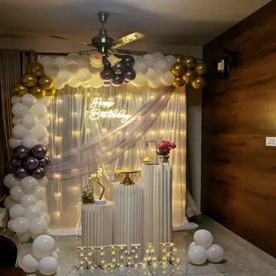 An elegant birthday party setup at home, featuring a white and gold balloon arch, a sheer curtain backdrop with fairy lights, and illuminated marquee letters spelling out the name 'KUMAR'.