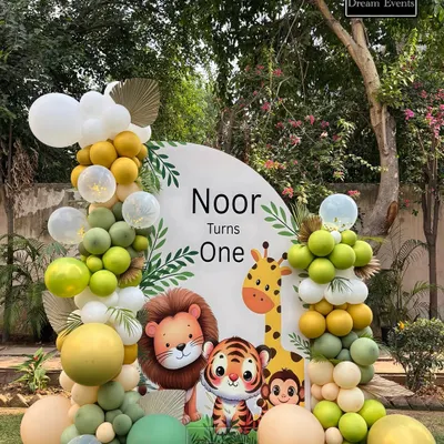 A custom circular backdrop for Noor's first birthday, featuring adorable safari animals like a lion, tiger, and giraffe. The theme is completed with organic balloon garlands in shades of green, yellow, and white.