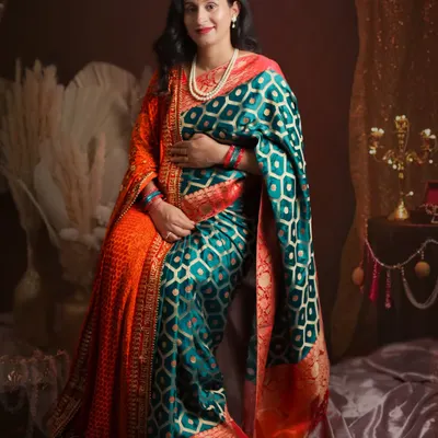 A full-length portrait of the mother-to-be in a stunning teal and orange saree. The studio setup includes pampas grass and traditional decor to complement the ethnic elegance of the attire.