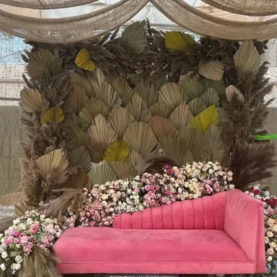 An elegant boho setup for a pre-wedding event, featuring a backdrop of dried palm leaves and a plush pink chaise lounge surrounded by flowers.