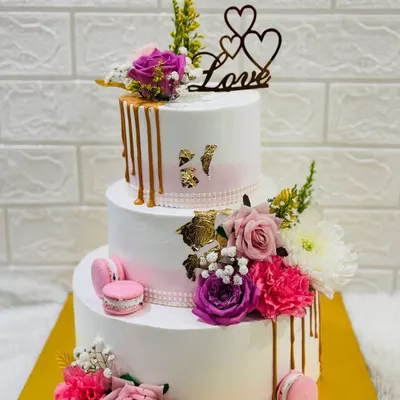 A magnificent three-tiered anniversary cake adorned with a cascade of fresh flowers, macarons, and a golden drip effect. A true centerpiece for a grand celebration of love.