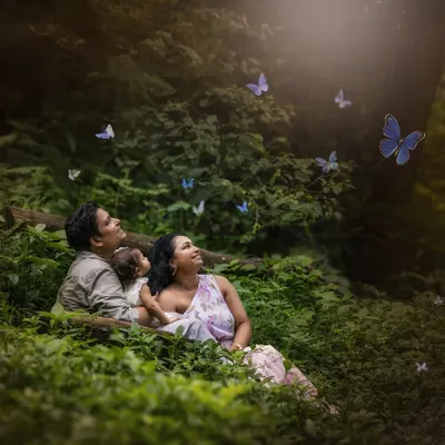 A family of three lies in a lush green forest, looking up in wonder as magical blue butterflies flutter around them. This composite image creates a dreamy, fairytale-like atmosphere.