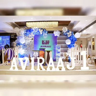 A cool and stylish stage for Aviraaj's Boss Baby first birthday. The setup includes a large "1" and the birthday boy's name in marquee letters, a hot air balloon prop, and a blue and silver balloon arch against a modern backdrop.