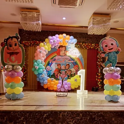A full-stage Cocomelon theme for a first birthday party. This setup includes a large circular backdrop with characters, rainbow balloon arch, light-up number '1', and character standees on balloon pillars.