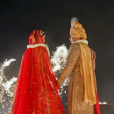 A classic portrait of the bride and groom in their wedding attire, holding hands against a backdrop of celebratory fireworks. A moment of quiet connection amidst the grandeur.
