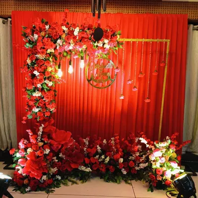 A vibrant and traditional backdrop for a pre-wedding ceremony. This setup features a rich red drape, a lush arch of red and white roses, and a custom gold acrylic sign with the couple's names.