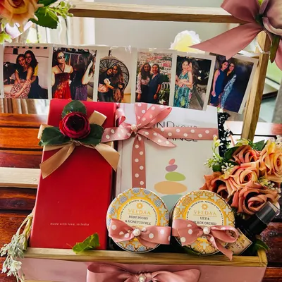 A close-up of a personalized hamper created for a best friend's wedding. The highlight is a string of cherished photographs, complemented by VILASA luxury wellness products and a gift box, all arranged in a beautiful wooden crate with pink floral details.