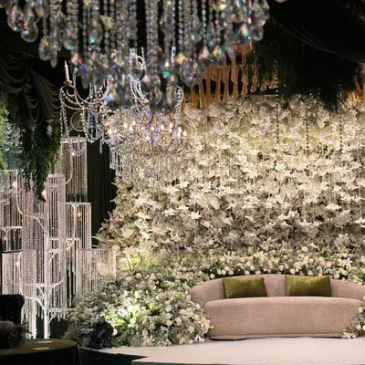 A full view of the enchanted forest stage, where a massive wall of white flowers serves as the backdrop. Crystal chandeliers and candle installations add layers of light and luxury to the magical setting.