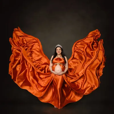 A breathtaking solo portrait using fabric tossing to create a dynamic, wing-like effect with a fiery orange satin gown. This technique adds movement and drama, making for a powerful and artistic maternity photo against a simple dark background.