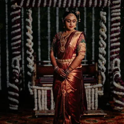 A full view of the traditional Tamil Brahmin bridal look, featuring the 9-yard Madisar saree. The rich maroon silk and intricate jewelry come together for a look of pure elegance in front of the Oonjal.
